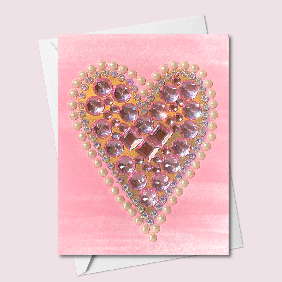 Pink greeting card with a gold-toned heart filled with pink jewels and edged in pearl-style embellishments, shown with a white envelope.