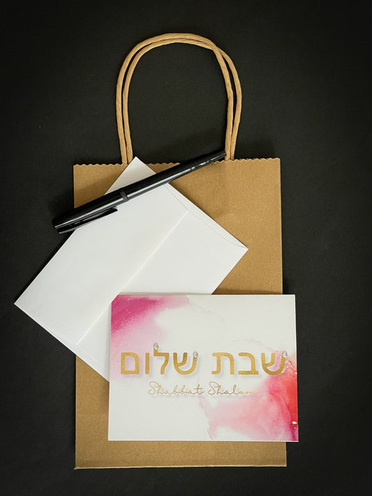 this is a note card on a craft paper gift bag with a white envelope and a black marker , the card says shabbat shalom in English and Hebrew with some pink watercolour accents