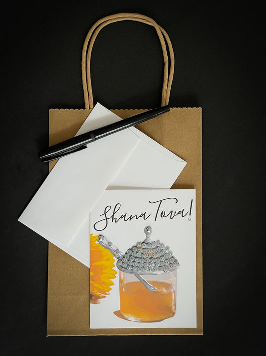 this is a note card on a craft paper gift bag with a white envelope and a black marker , the card says Shana Tova with a a jar of honey