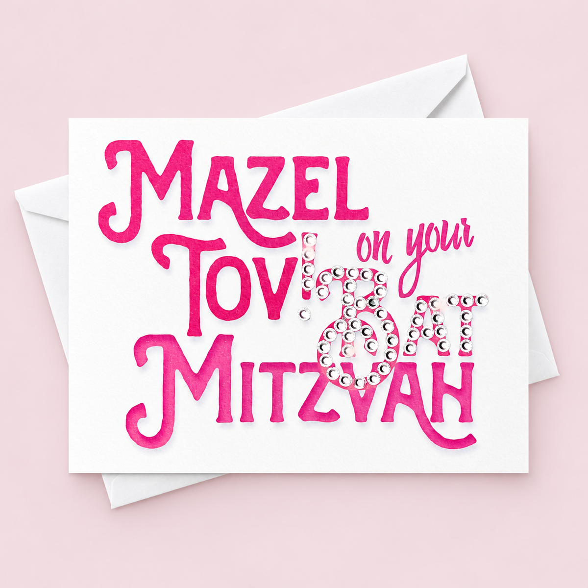 Mazel Tov on your Bat Mitzvah card with pink lettering and crystal embellishments, with a white envelope on a pale pink background.