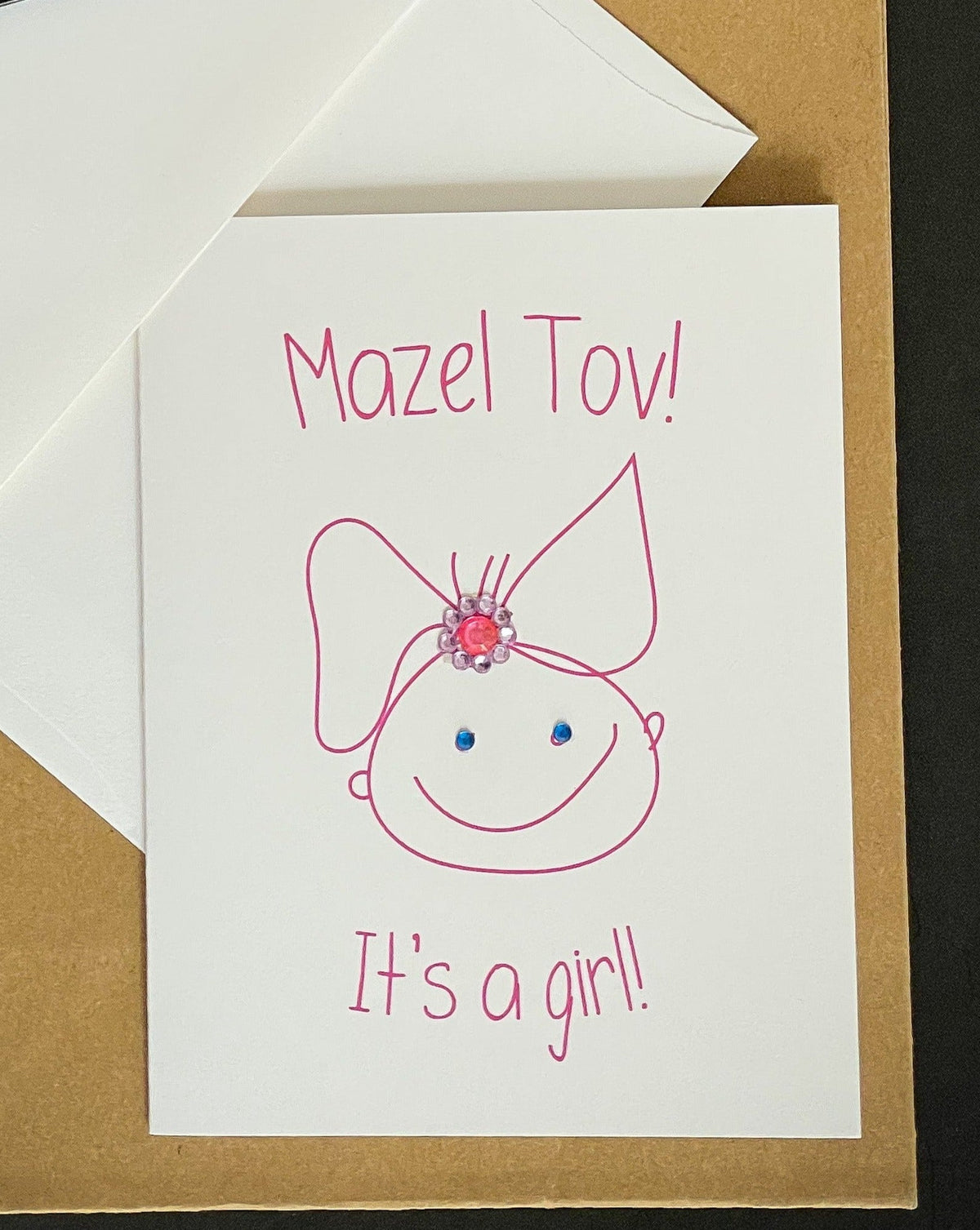 Original photograph of the Mazel Tov! It’s a Girl! card with pink lettering, baby-girl illustration and jewel details, on kraft paper with a white envelope.