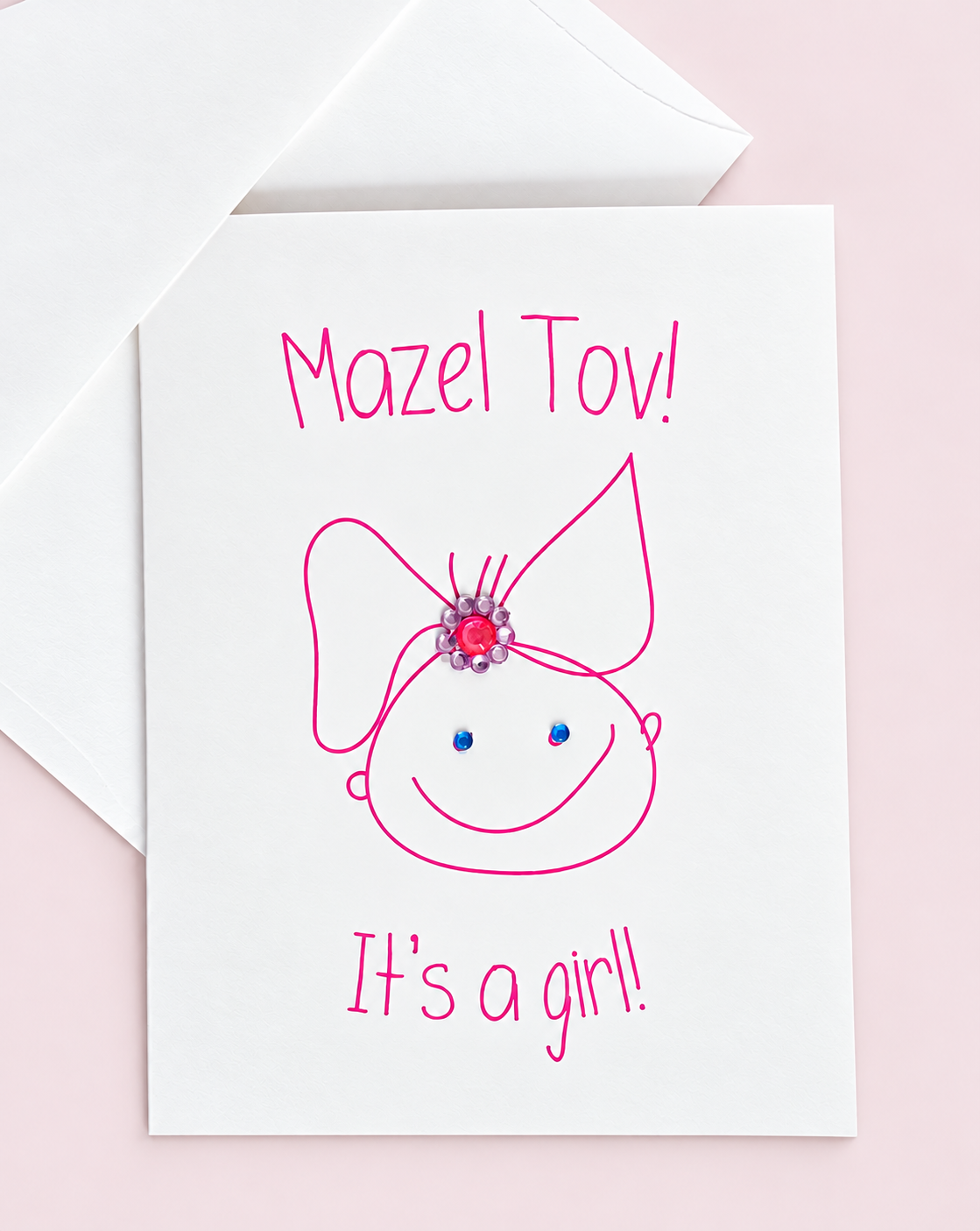 Mazel Tov! It’s a Girl! greeting card with a pink baby-girl illustration and jewel embellishments, pictured with a white envelope on a pale pink background.