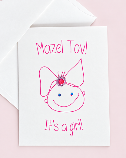 Mazel Tov! It’s a Girl! greeting card with a pink baby-girl illustration and jewel embellishments, pictured with a white envelope on a pale pink background.