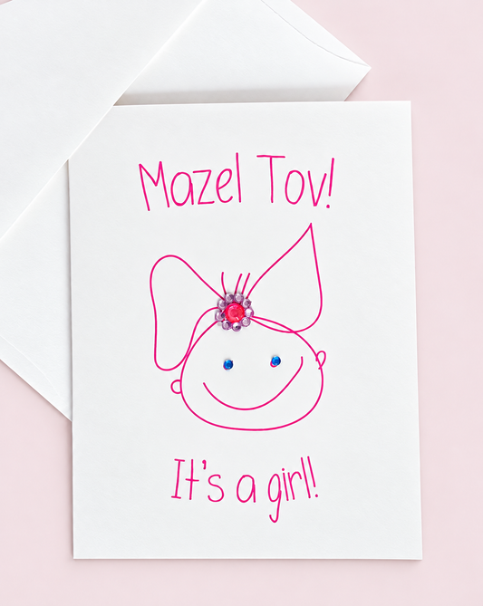 Mazel Tov! It’s a Girl! greeting card with a pink baby-girl illustration and jewel embellishments, pictured with a white envelope on a pale pink background.